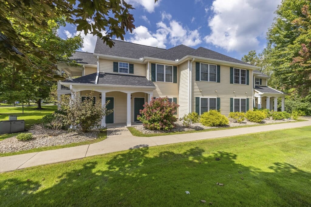 Two-story beige house with green shutters, surrounded by landscaped shrubs and trees, under a blue sky with clouds.