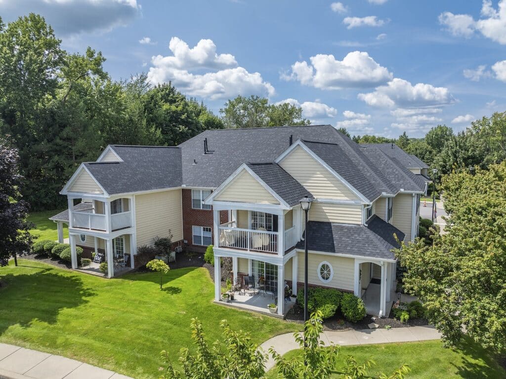 An aerial view of a suburban apartment complex with attached balconies, surrounded by green lawns and trees under a partly cloudy sky.