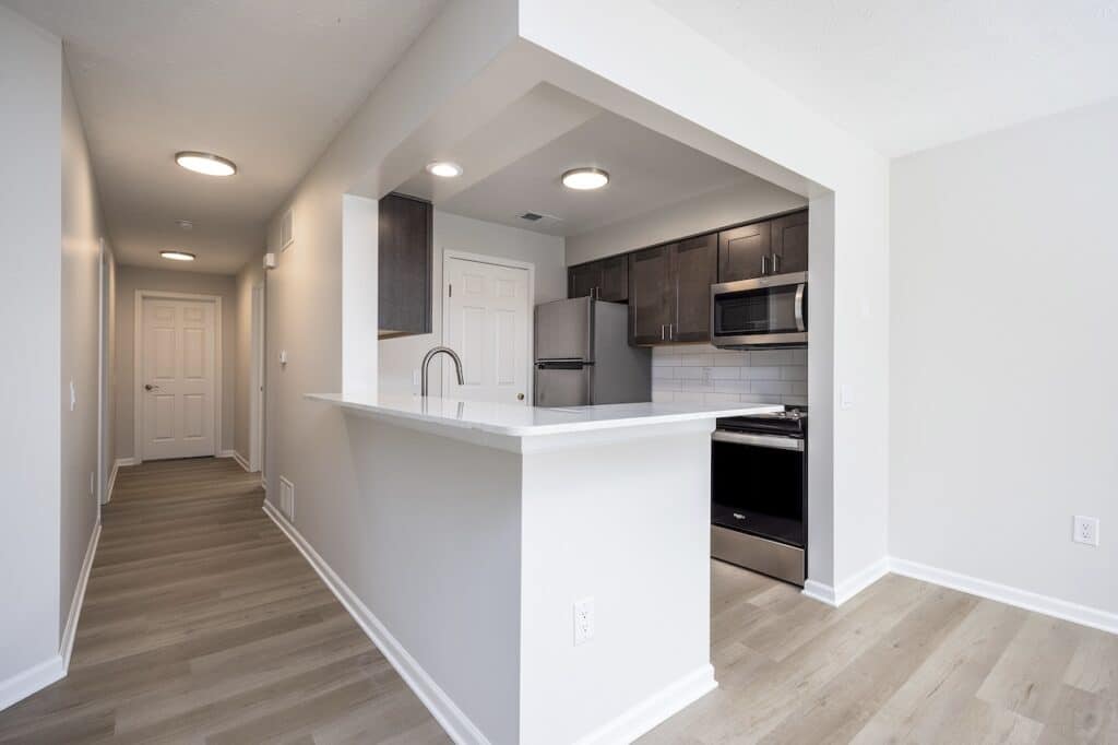 Modern kitchen with dark cabinets, stainless steel appliances, and white countertops. Adjacent hallway with light wood flooring and three closed doors.