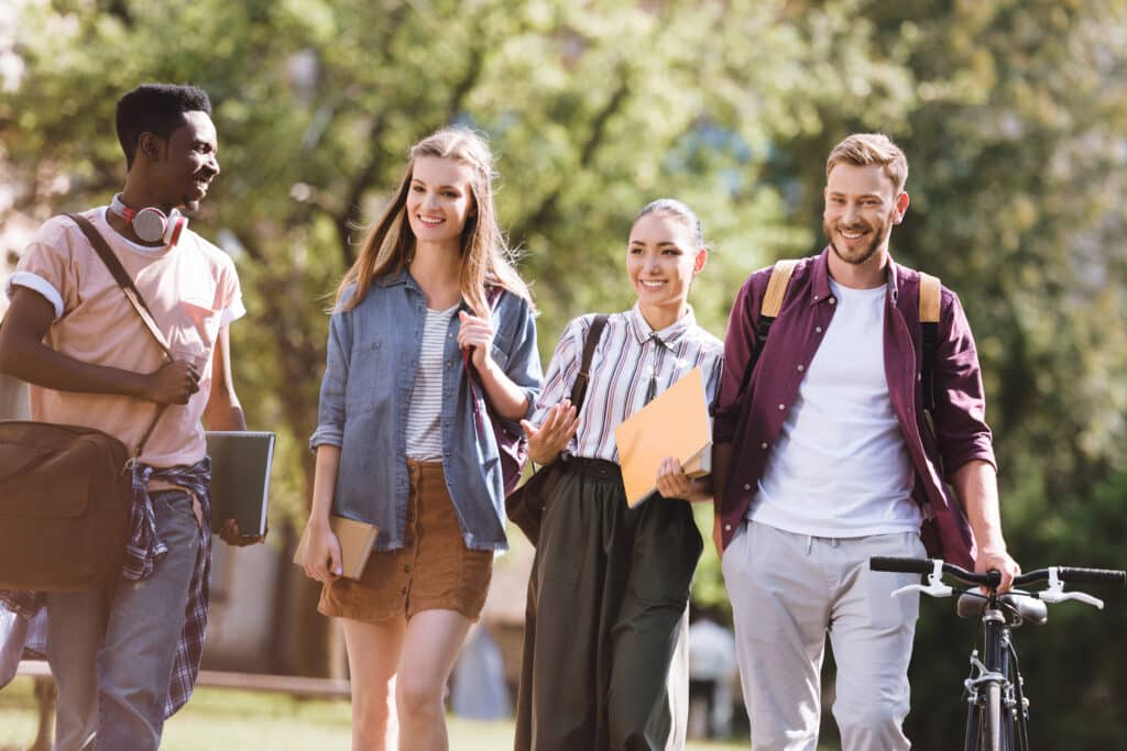 Four young adults walking outdoors, carrying books and a bicycle, smiling and engaging in conversation.