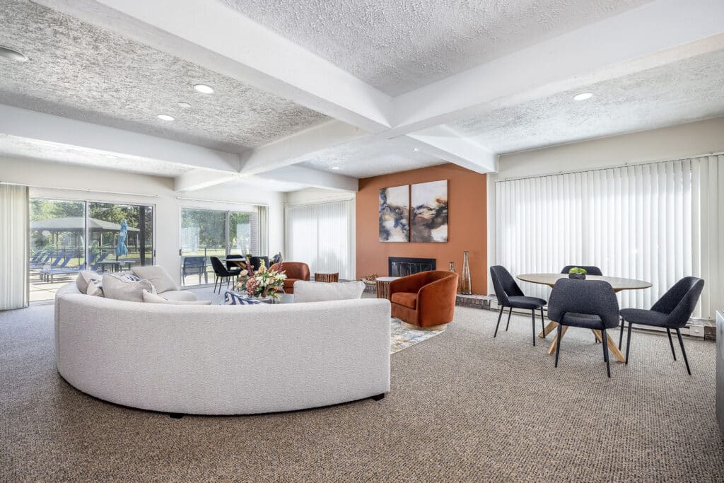 Spacious living room with a curved white sofa, armchairs, round dining table with chairs, and a fireplace. Large windows and a glass door offer views of a patio and swimming pool.