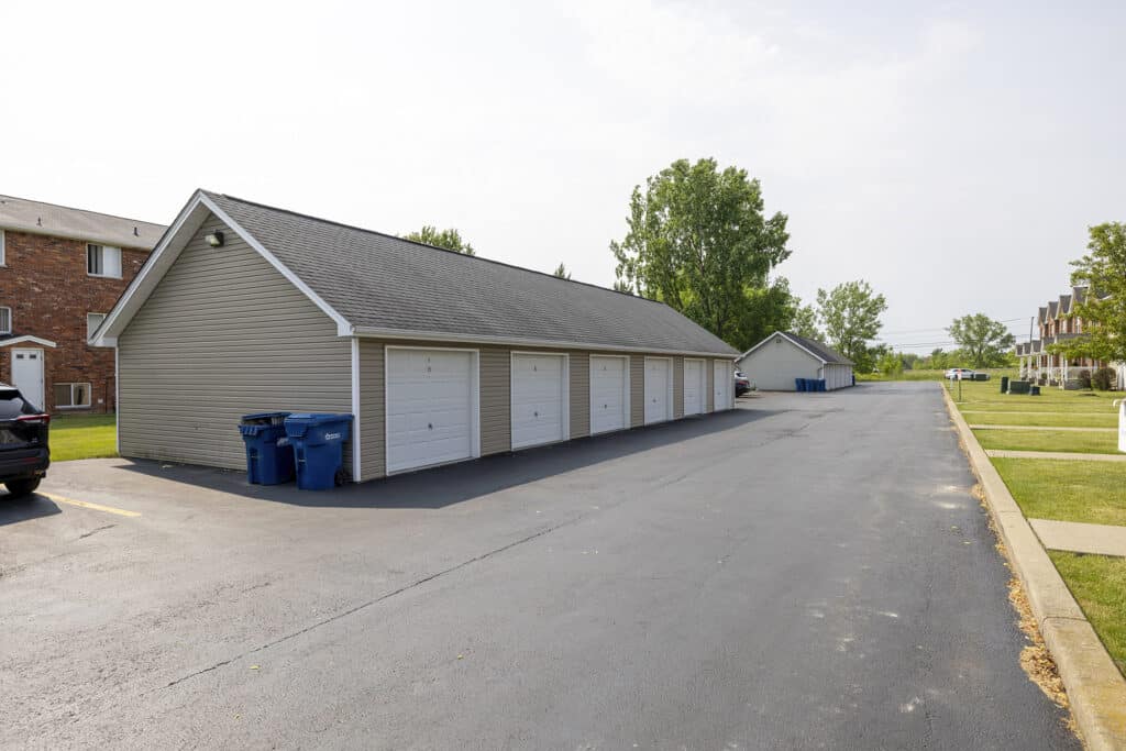 A row of garages with white doors and gray siding in a suburban parking lot, with several recycling bins nearby.