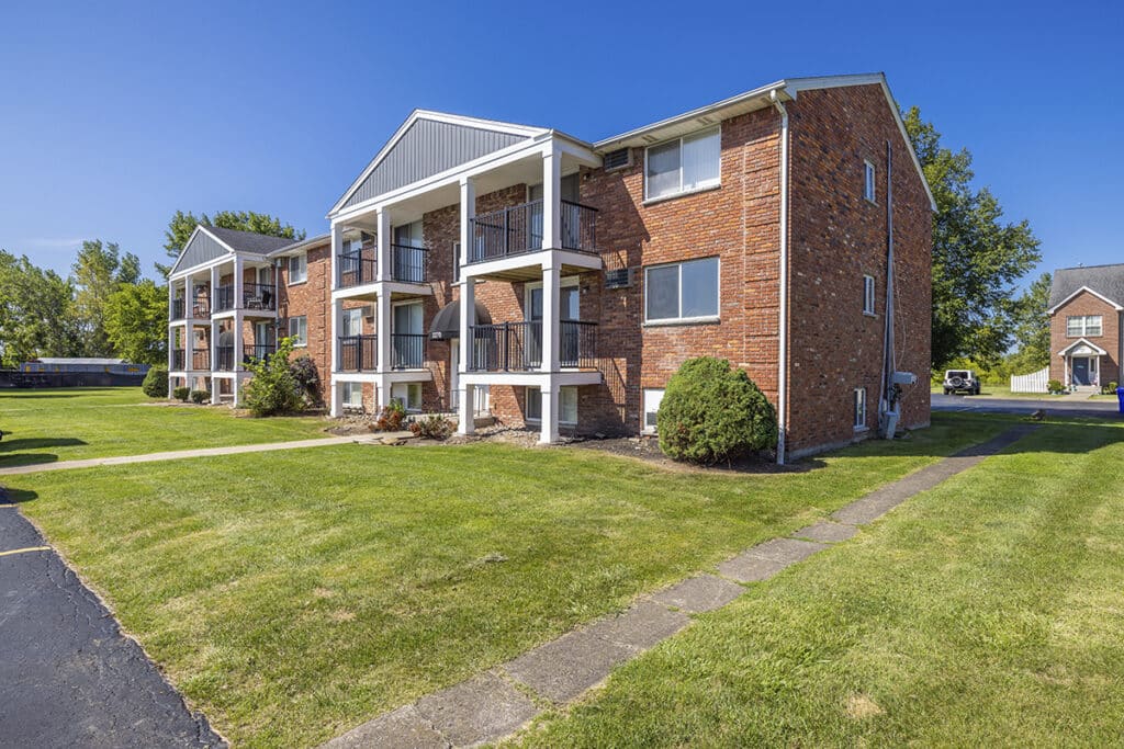 A three-story brick apartment building with white balconies, surrounded by green lawns and trees under a clear blue sky.