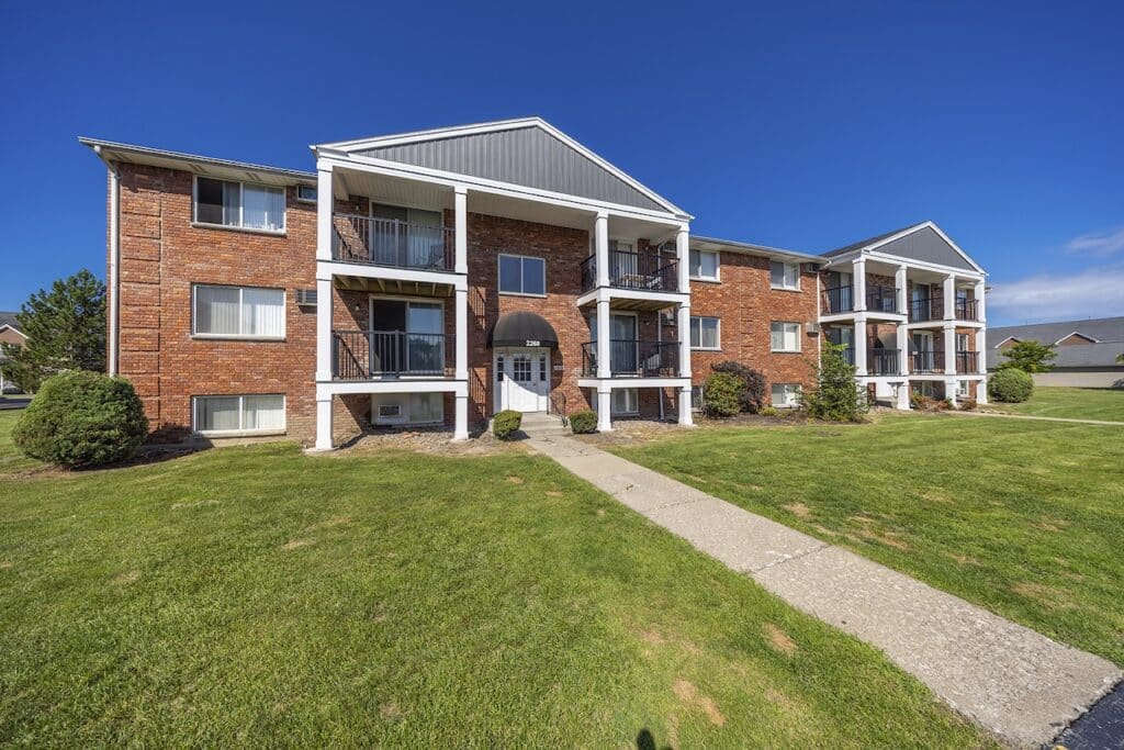 A two-story brick apartment building with balconies, surrounded by a grassy lawn under a clear blue sky.