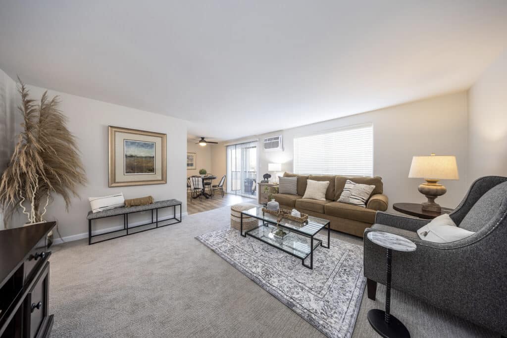 A spacious living room with a brown sofa, glass coffee table, and gray armchair. A painting and pampas grass adorn the walls. The room is well-lit with a lamp and natural light from a window.