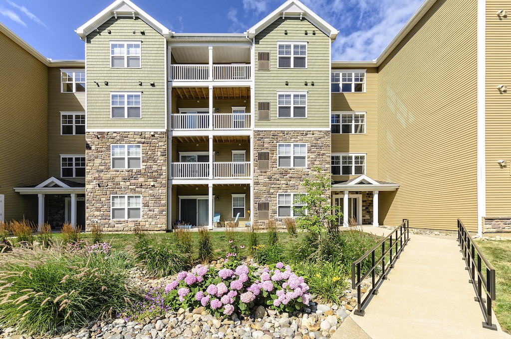 Four-story residential building with balconies, tan and green siding, and stone accents. A garden with pink hydrangeas and a paved walkway are in the foreground under a blue sky.