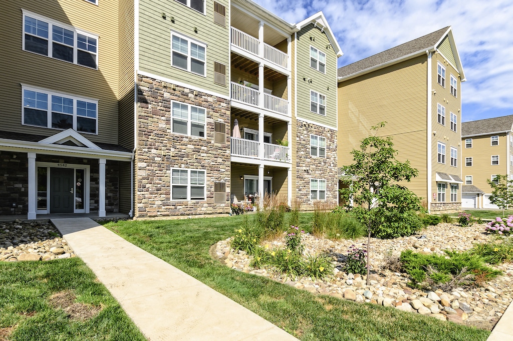 Four-story residential apartment building with stone and siding facade, balconies, and a landscaped garden. A stone path leads to the entrance.