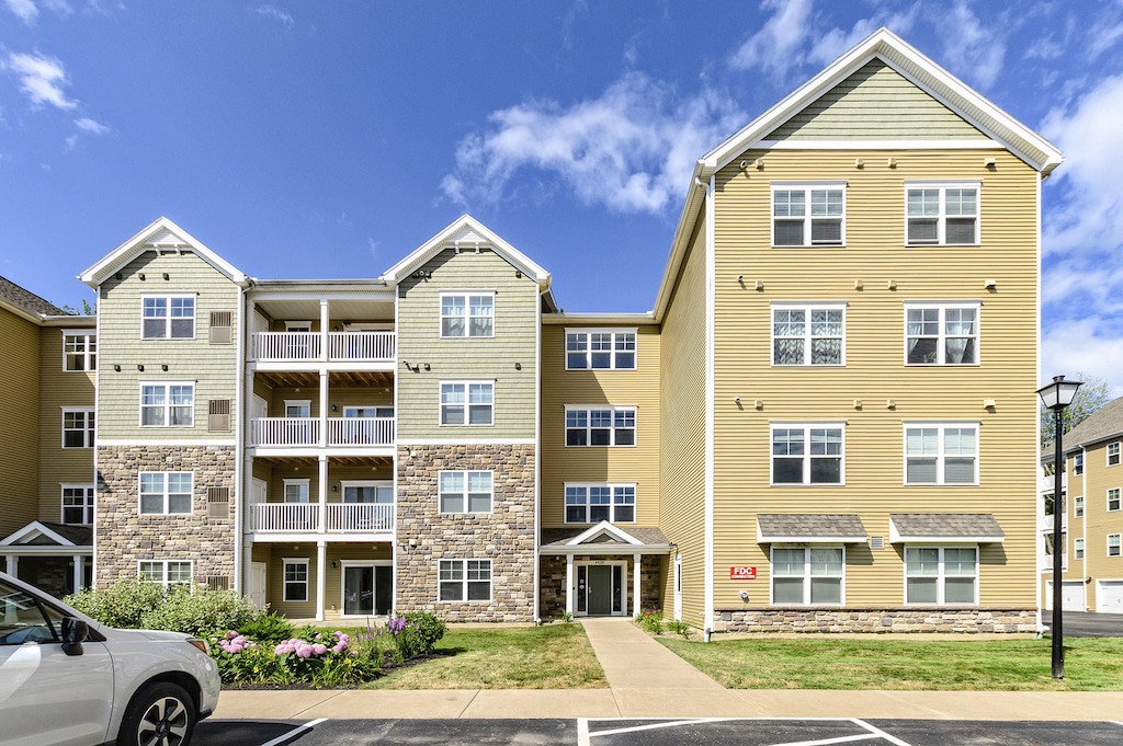 A four-story apartment building with beige siding and a stone facade, featuring balconies and a sidewalk leading to the entrance, set under a blue sky with clouds.