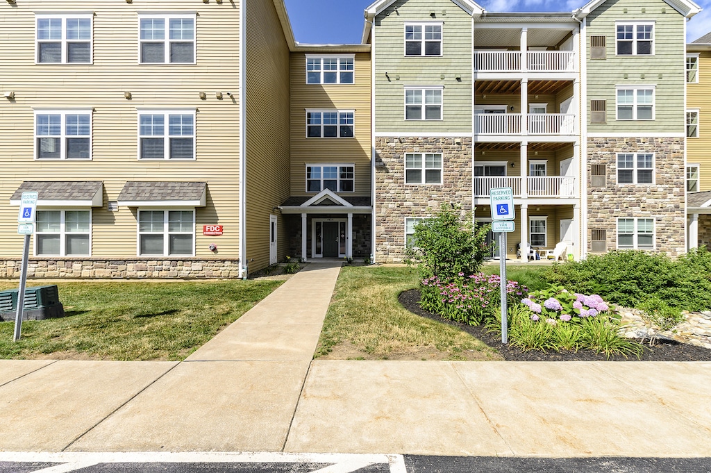 Three-story apartment building with beige and green siding, stone accents, balconies, and landscaped front with flowers. Sidewalk and parking spaces in foreground.