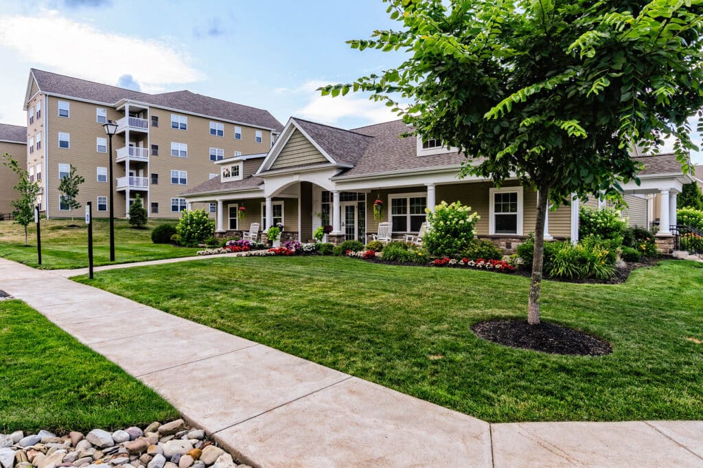 Wide view of a landscaped residential area with a pathway, a one-story house surrounded by greenery, and a multi-story building in the background.