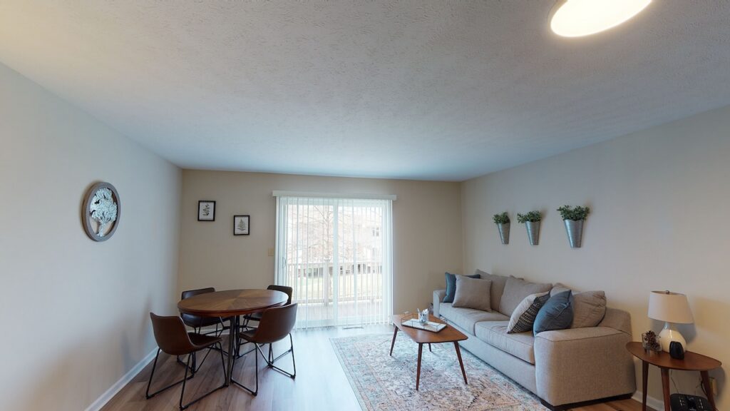 A living room with a gray sofa, a round wooden table with four chairs, wall-mounted plants, and a rug. Sliding glass doors lead to a balcony, allowing natural light to fill the space.