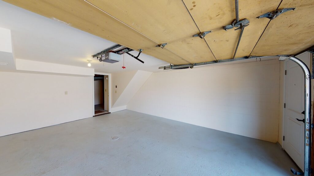 Empty garage interior with closed wooden ceiling door, white walls, and concrete floor. A single light bulb is on the ceiling, and a side door is partially visible to the right.