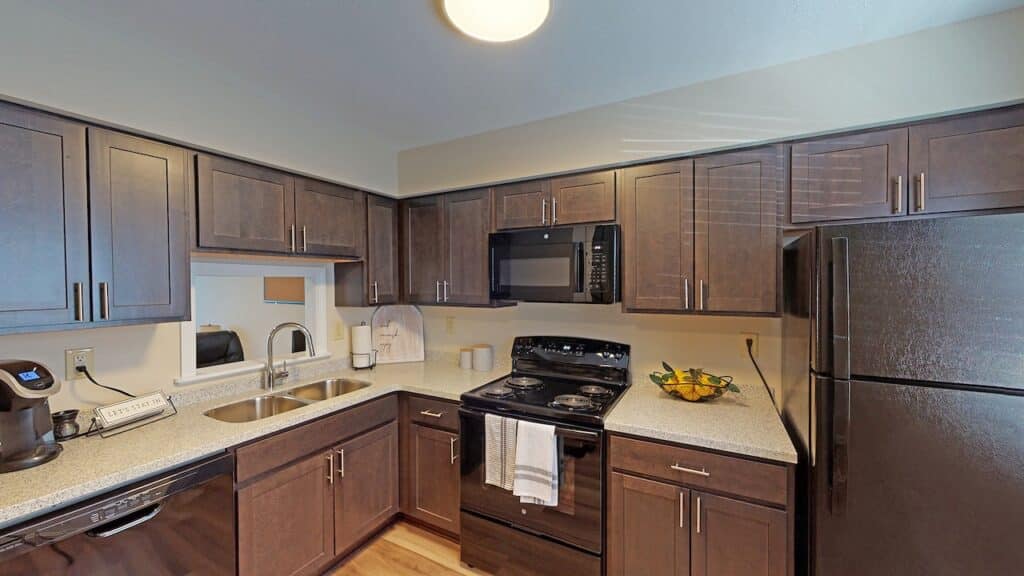 Modern kitchen with dark wood cabinets, granite countertops, black appliances, and a double sink. A towel hangs on the oven door, and a bowl of fruit sits on the counter.