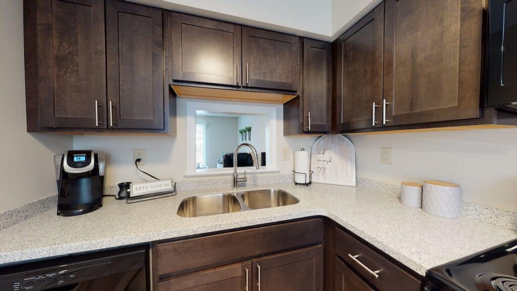 Modern kitchen with dark wood cabinets, a dual sink, a coffee maker, and countertop decor. A small window opens to another room.