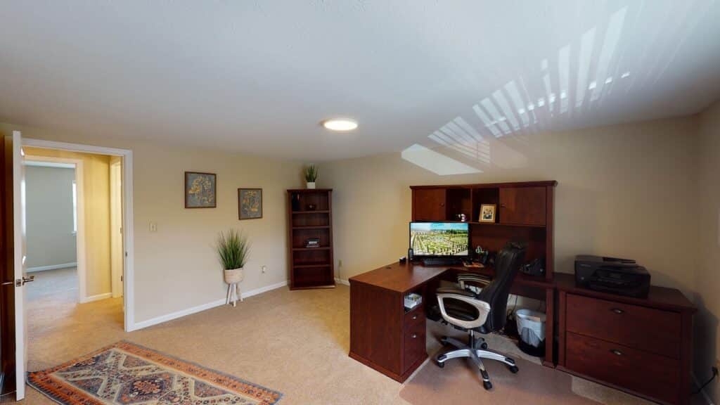 Home office with a wooden desk, office chair, computer, and printer. Bookshelf and framed pictures on the wall. Beige carpet and an open door leading to another room.