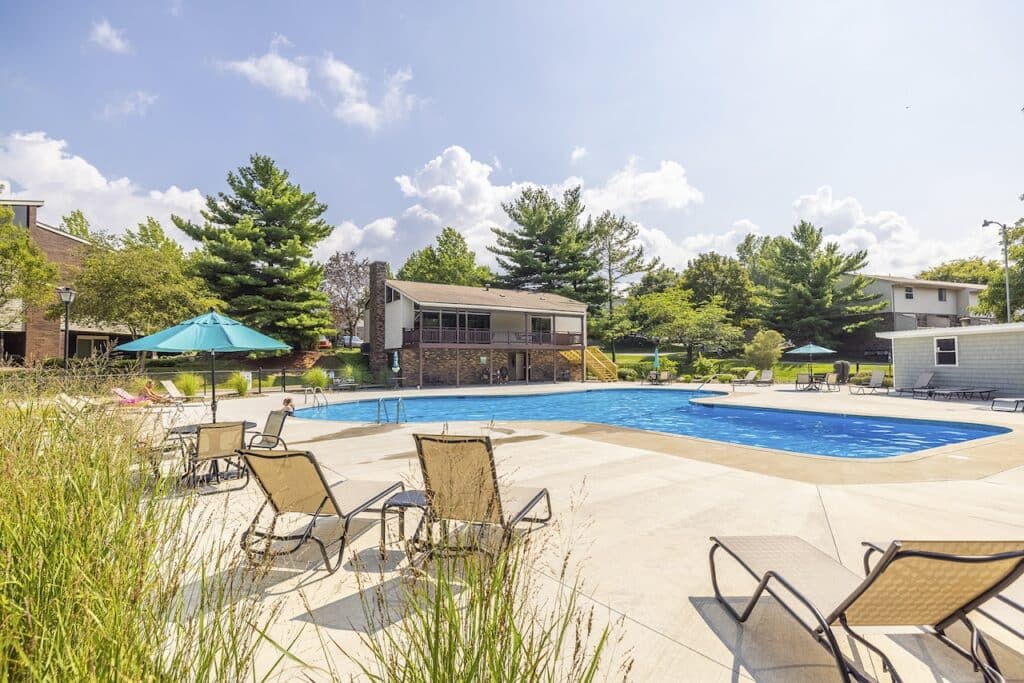 Outdoor pool area with several lounge chairs, umbrellas, and surrounding trees. A small building is visible in the background.