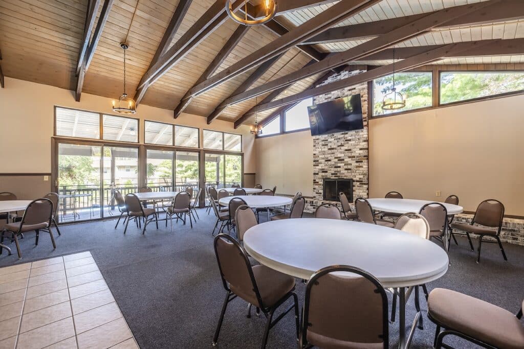 A spacious room with round tables and brown chairs, featuring a high wooden ceiling, large windows, and a stone fireplace with a mounted TV. Natural light streams through the windows.
