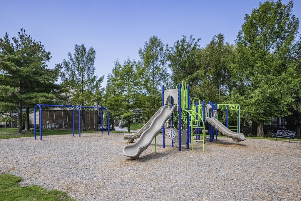 Playground with slides, swings, and climbing structures on a gravel surface, surrounded by trees under a clear sky.