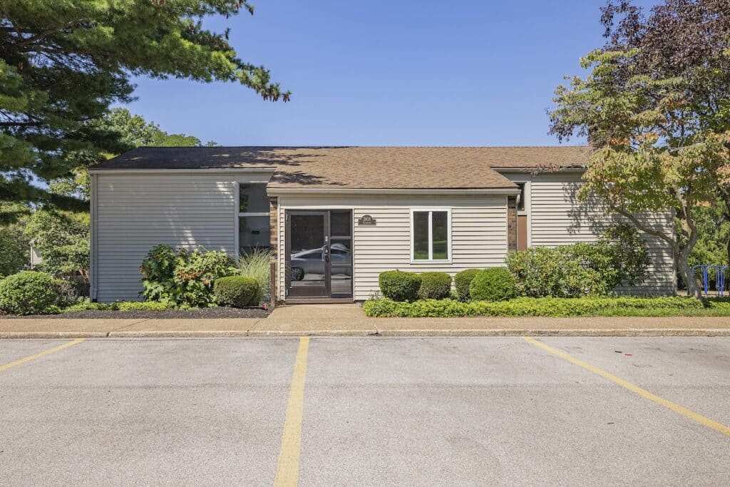 Single-story house with gray siding, a central front door, and a small garden. A paved driveway and parking space are in the foreground, with trees surrounding the property.