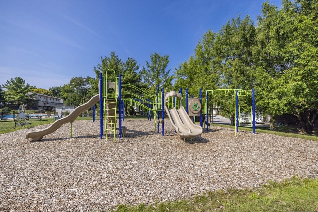 A playground with slides, climbing structures, monkey bars, and wood chip ground, surrounded by trees and grass under a clear blue sky.