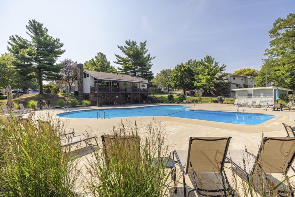 Outdoor swimming pool with lounge chairs, surrounded by trees and buildings. A wooden building with lifebuoys is in the background.