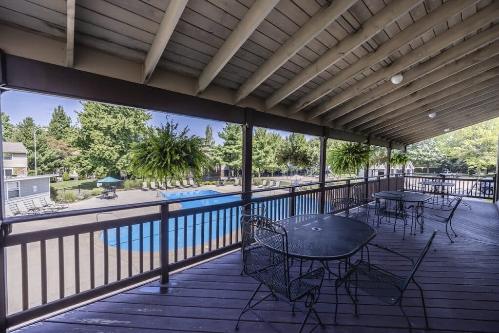 Covered patio with metal tables and chairs overlooking an outdoor pool surrounded by lounge chairs and trees.