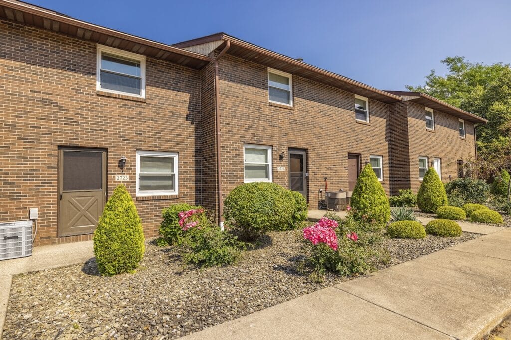 A row of two-story brick townhouses with manicured shrubs and flowering plants in front, set on a clear sunny day.