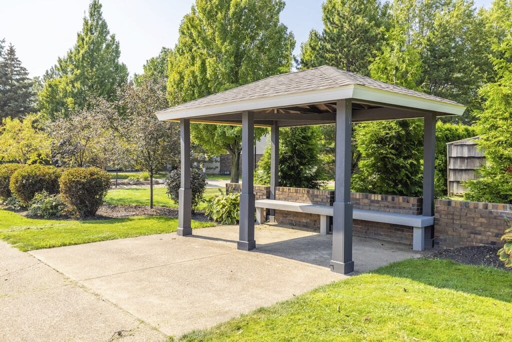 A small, open-sided gazebo with a gray roof and benches stands on a concrete platform surrounded by green trees and shrubs in a park setting.