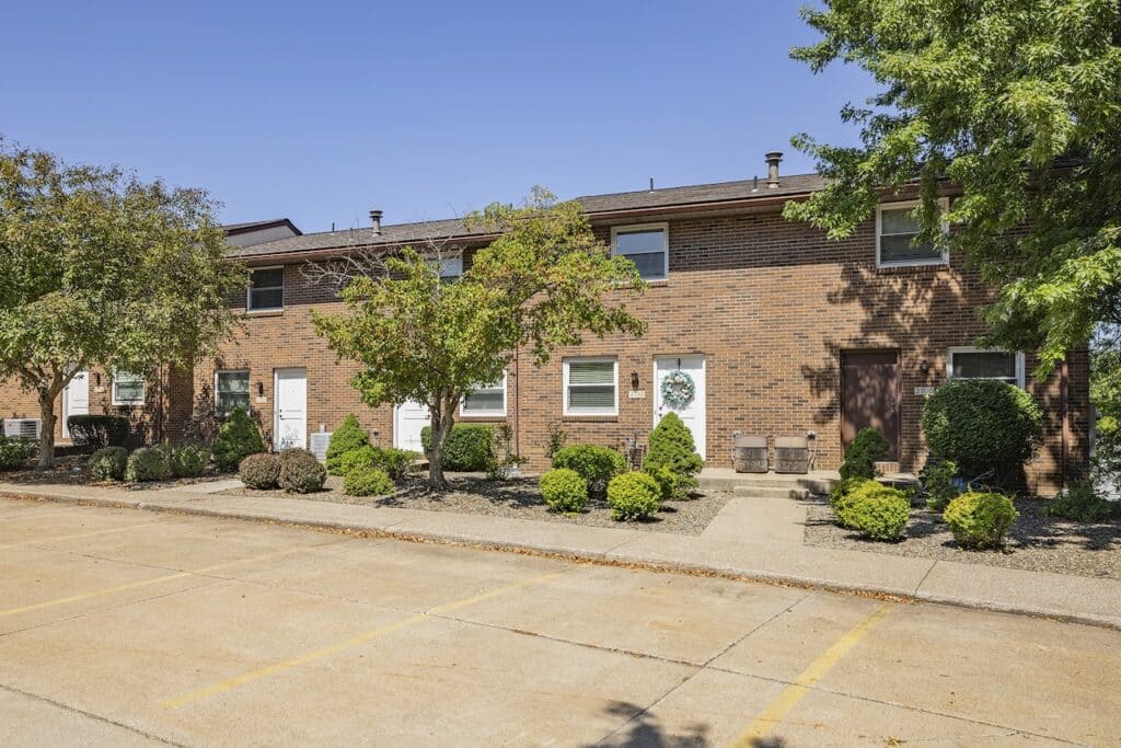 A row of brown brick townhouses with small shrubs and trees in front, a clear blue sky above, and an empty parking area in the foreground.