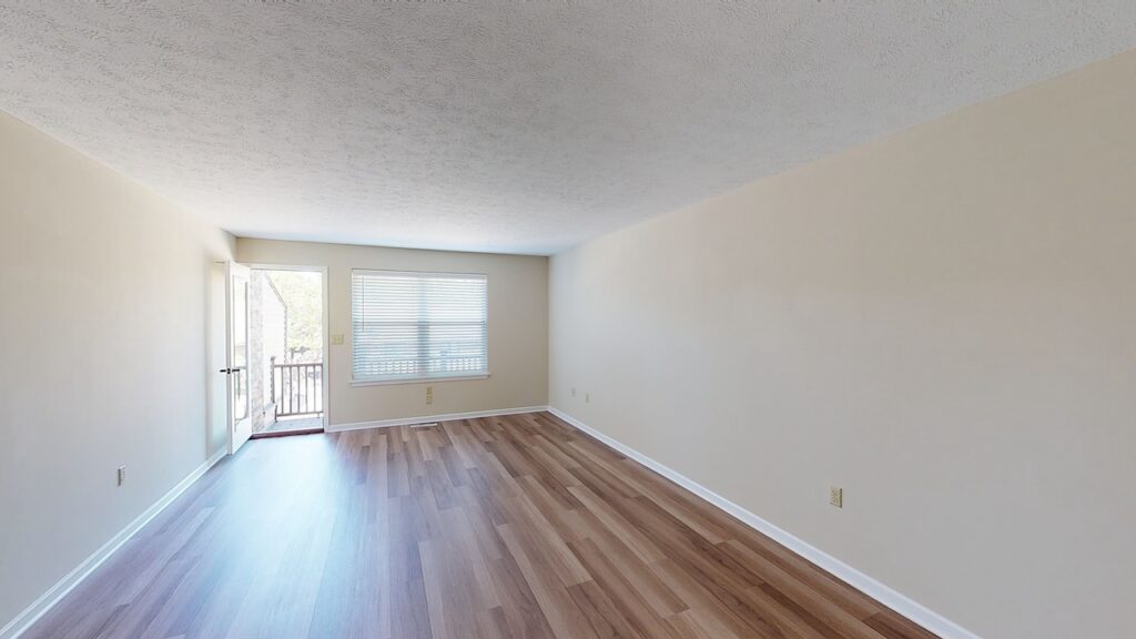 An empty room with light brown laminate flooring, beige walls, a large window with blinds, and a door leading to a balcony.
