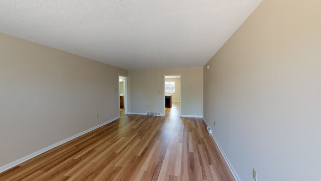 Empty room with beige walls and wooden flooring, leading to another room with a doorway. Natural light enters from a window in the background.