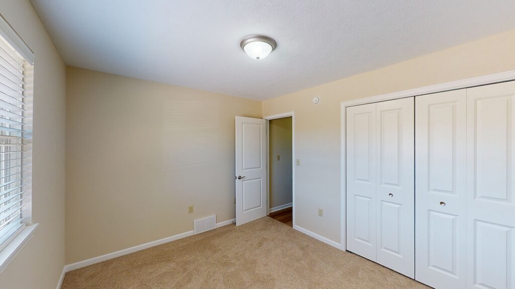Empty room with beige walls, a ceiling light, carpeted floor, open door, and closed double-door closet. Sunlight enters through a window with blinds on the left.
