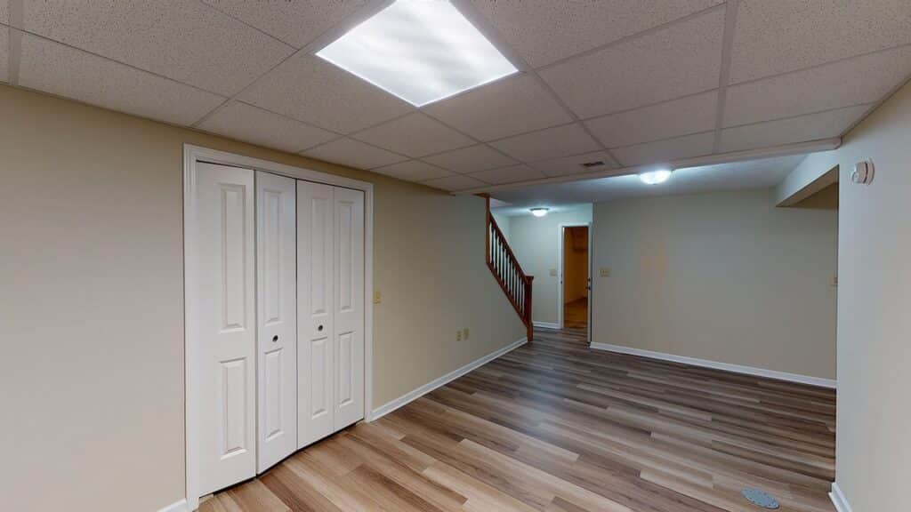 Basement room with beige walls, wood laminate flooring, closed white double doors, a staircase, and ceiling lights.