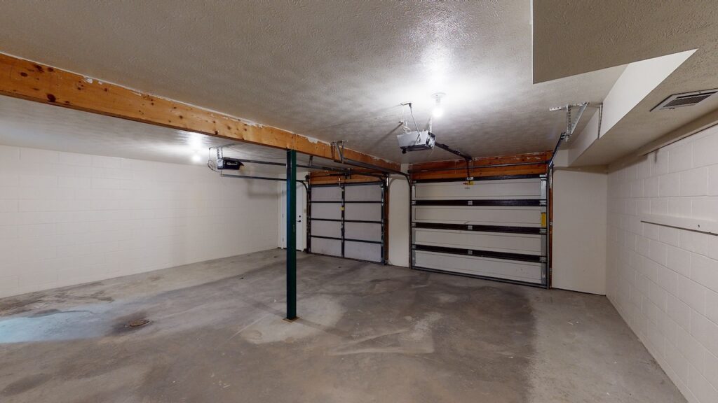 Empty garage with concrete floor, two closed garage doors, and overhead lighting. Wood beam supports ceiling.