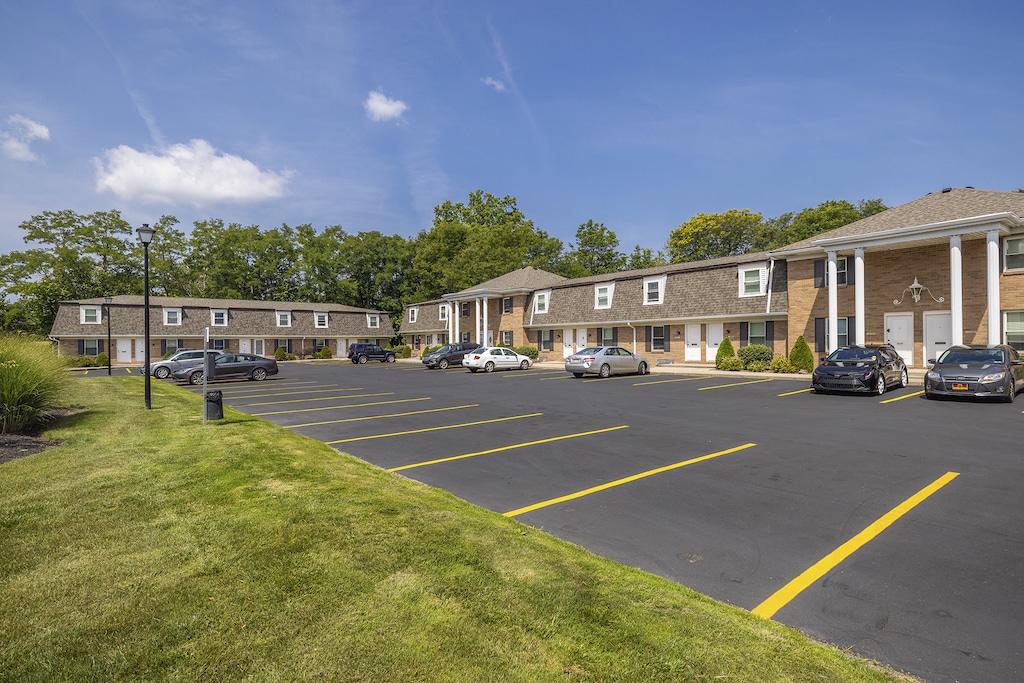 Apartment complex with two-story buildings and a parking lot, featuring several parked cars. The sky is clear and sunny.