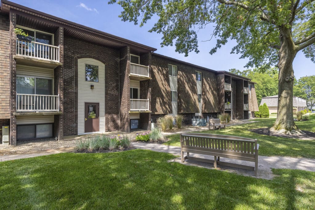 A three-story brick apartment building with balconies and surrounding trees, grassy lawn, and a bench in the foreground.