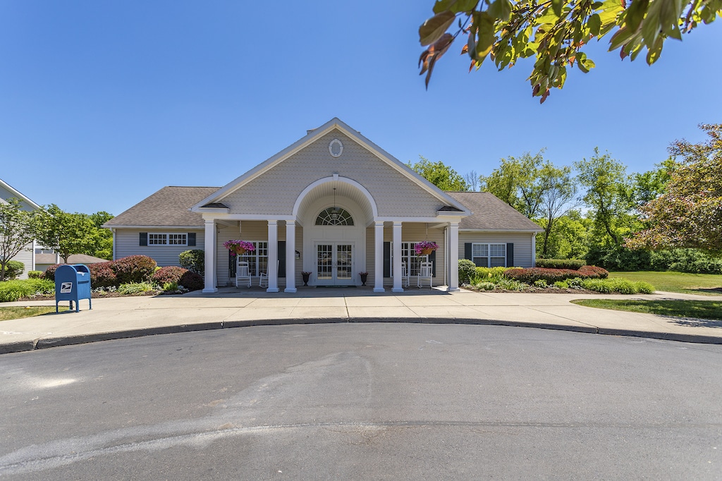 A single-story building with a circular driveway, featuring a gabled roof, columns at the entrance, and a mailbox to the left. Surrounded by greenery under a clear blue sky.