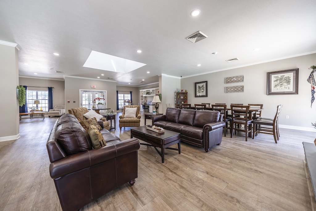 A spacious living room with brown leather sofas, armchairs, a dining table, and wooden flooring. The room has large windows, wall art, and overhead lighting.