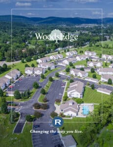 Aerial view of a residential community with multiple white buildings, a parking lot, and a swimming pool, surrounded by greenery and distant hills. Text reads "WoodsEdge" and "Changing the way you live.