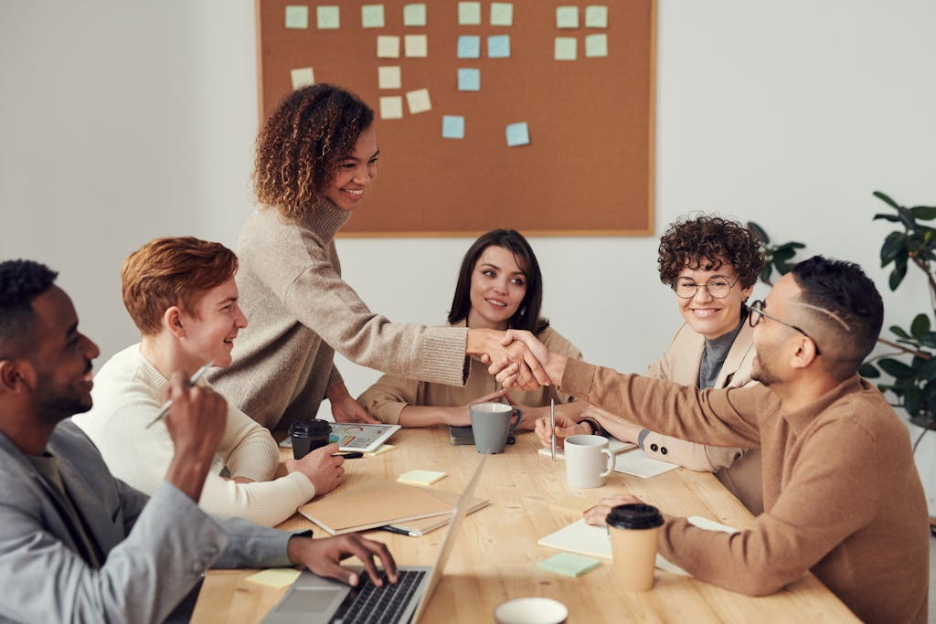 A group of people seated at a table in a meeting room, with two individuals shaking hands. Laptops, coffee cups, and a corkboard with sticky notes are visible in the background.