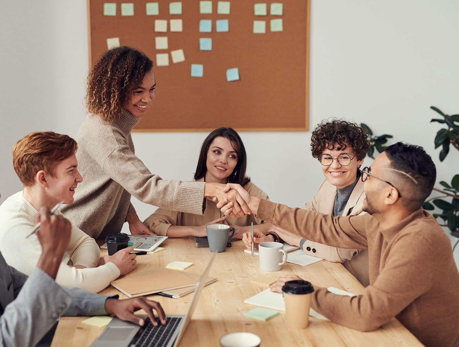 Five people in a meeting room, with one person shaking hands with another. Laptops, coffee cups, and sticky notes are visible on the table and wall.