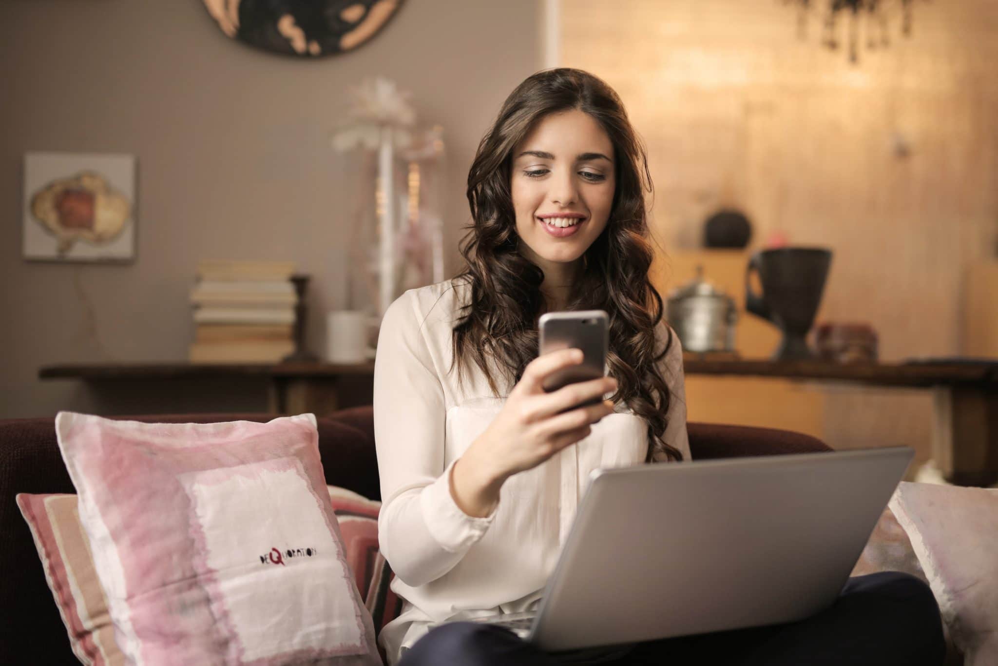 A woman sitting on a couch uses a phone and laptop simultaneously, with a cozy living room setting in the background.