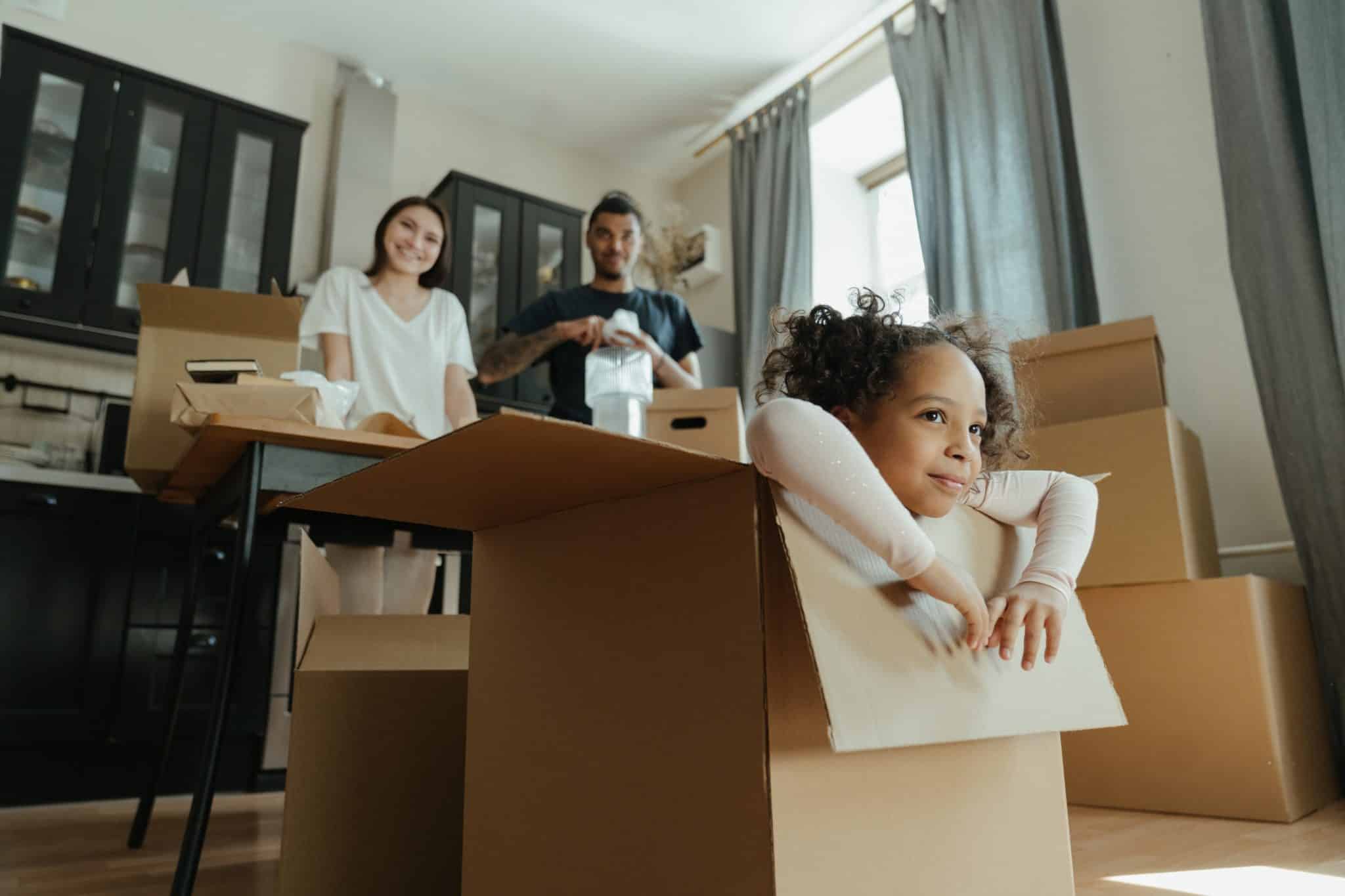 Child sitting in a cardboard box in a room with two adults and more boxes, suggesting a moving or unpacking scene.