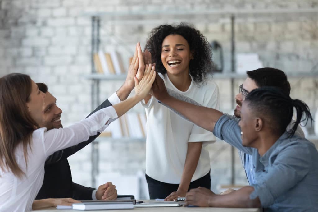 A diverse group of five people smiling and high-fiving around a table in an office setting.