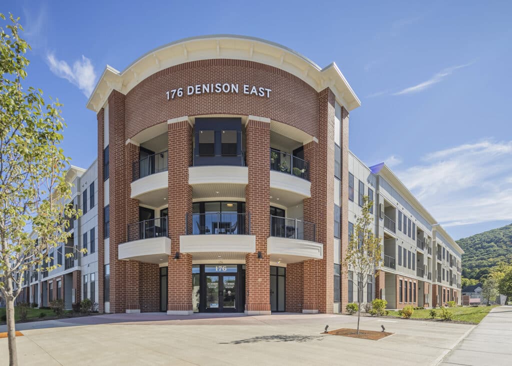 Three-story brick building with white accents and columns, labeled "176 Denison East." Surrounded by trees and a clear blue sky.