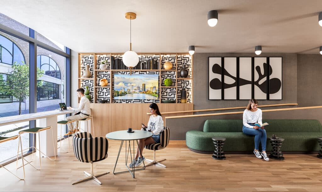 A modern cafe with three people. One reads on a green sofa, another drinks by a round table, and the third gazes out a window from a high stool. Art and greenery decorate the space.