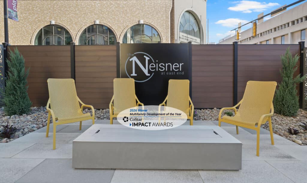 Four yellow chairs surround a rectangular fire pit on a patio at The Neisner at East End, in front of a sign and an award plaque for Multifamily Development of the Year.
