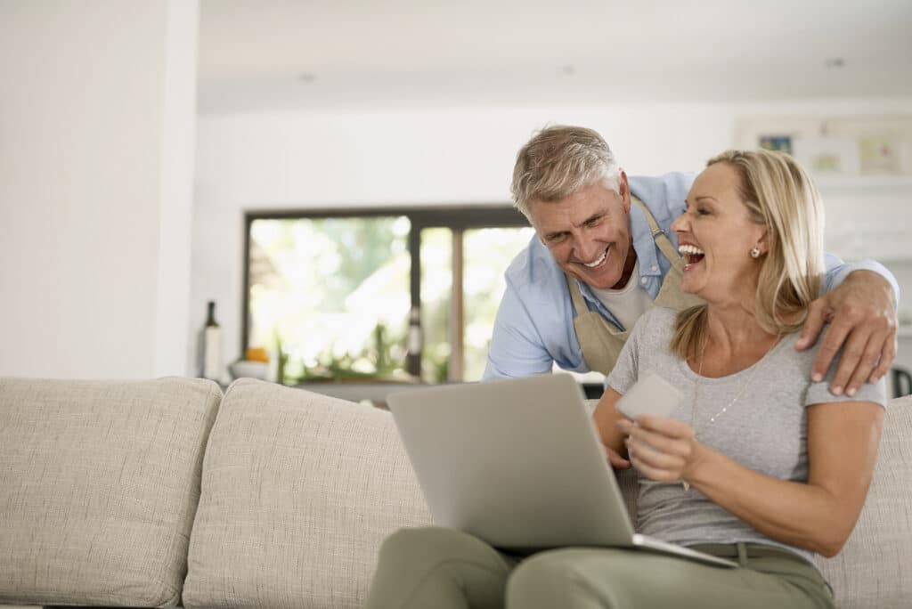 A couple sits on a couch, smiling and looking at a laptop. The woman holds a card in her hand.