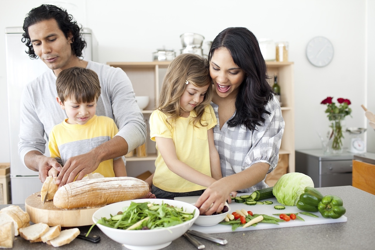 A family of four prepares a meal together in a kitchen. An adult slices bread, while another assists children with vegetables. A salad bowl is in the foreground.