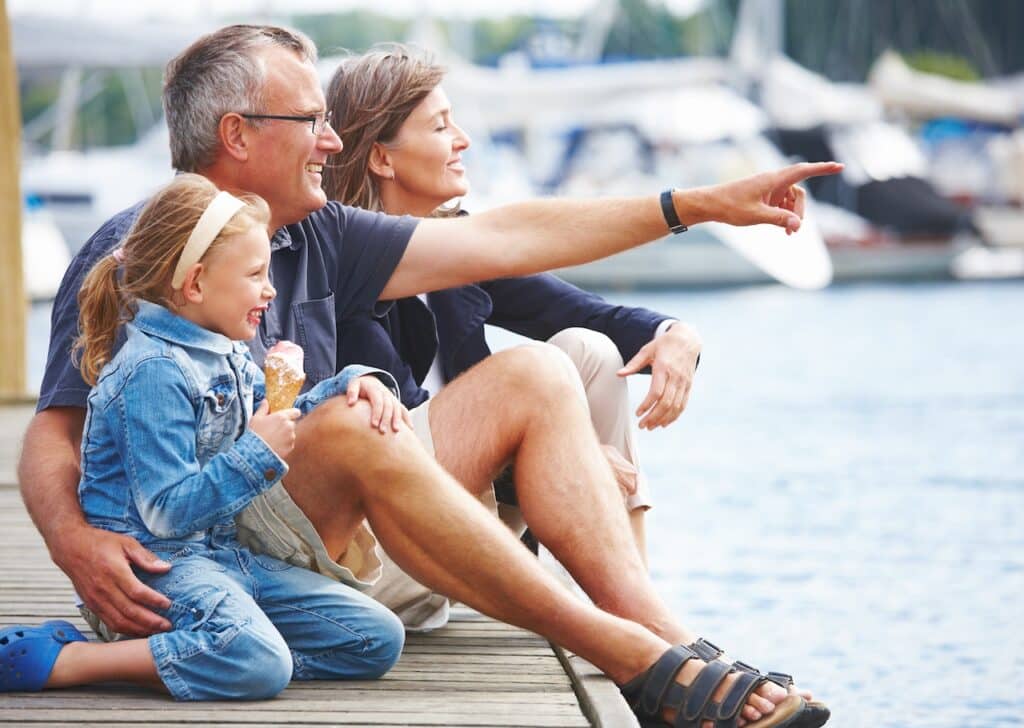 A family of three sits on a dock by the water. The man points into the distance while the woman smiles, and the young girl eats an ice cream.